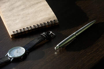A wrist watch, a ballpoint pen and a notebook with a spring lie on a dark wooden table. Concept of creative work, testament, memoir and deadline. Focus stacking