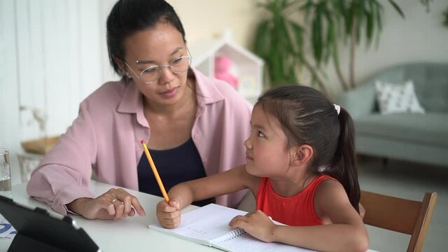 Online distance: woman helping girl to study and sitting at table in front of tablet in room spbd. Young mother teaches daughter and talks, cute girl writes in notebook and looks at device screen