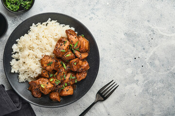 Spicy teriyaki chicken fillet pieces with rice, green onions and black sesame seeds on black plate on a grey slate, stone or concrete background. Top view with copy space.
