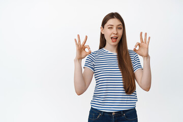 Young brunette woman winking, showing okay signs in approval, making compliment, standing over...