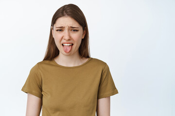 Young woman stick tongue from dislike, disgusted with bad taste, smth awful or disgusting, standing over white background - obrazy, fototapety, plakaty