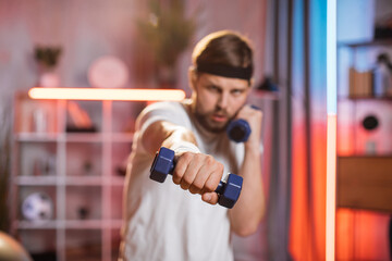 Fototapeta na wymiar Front blurred view of likable concentred active sporty man in sportswear, standing in front of camera at home and making boxing punches with dumbbells. Focus on hand with blue dumbbell