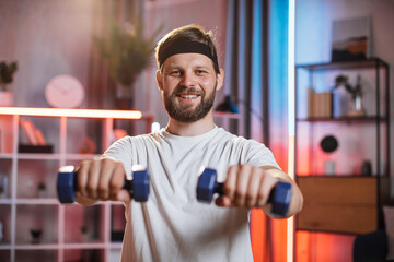 Smiling caucasian man in headband using dumbbells for workout at living room. Happy bearded guy...