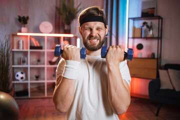 Fototapeta na wymiar Middle aged with vivid facial expression lifting dumbbells during evening time at home. Portrait of male beginner in headband making effort for training.