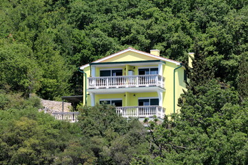 Fototapeta na wymiar New large yellow suburban family house with two elongated white front balconies and attached terrace built on side of small hill surrounded with dense trees on warm sunny spring day