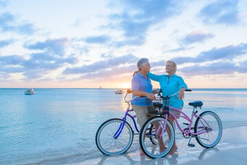 Fototapeta premium Older couple, seniors, riding bicycles on the beach in tropical blue outfits at sunset, romantic, fitness