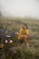 Woman traveler prepares breakfast, frying eggs on a burner while traveling in mountains on foggy weather