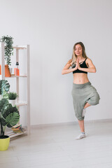 Woman practise yoga performs. Tree pose standing on mat inside of cozy room with plants and greenery. Healthy lifestyle concept