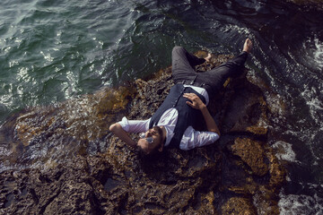 man with a beard in dark clothes and a white shirt lie on the stone seashore in Crimea tarkhankut