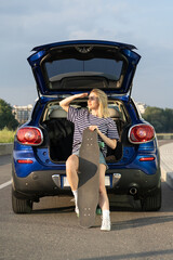 Woman sitting at back of car smiling enjoy sunset during summer vacation and adventure road trip. Middle aged female with skateboard travel in vehicle in summertime. Lifestyle and tourism concept