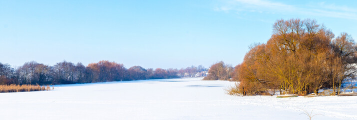 Winter landscape with trees on the banks of the river covered with ice and snow on a sunny day, panorama