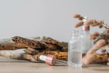 Transparent glass bottle with pipette, scincare serum or essential oil, dried flowers and old wood branch on wooden table. Natural organic cosmetics.