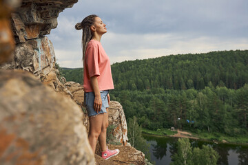 Naklejka premium a modern girl with dreadlocks and pink sneakers stands with her eyes closed at the entrance to the cave on top of the cliff, overlooking the lake and the dense forest. High quality photo