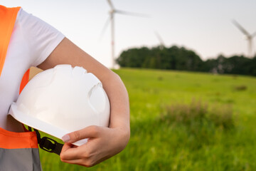 An engineer with a helmet in his hands and orange vesta stands on the background of windmill or...