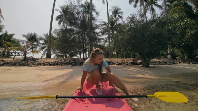 Young woman in sunglasses pulls pink plastic canoe with paddle to azure sea water from beach with palms. The beginning of the boat trip on kayak, boarding sailing. Traveling to tropical countries.