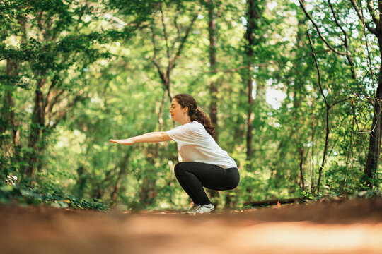 A young woman in sports clothes does a squat exercise. Summer forest in the background. Low angle. Side view. The concept of fitness and training in the park