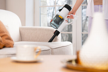 Young woman vacuums sofa with cordless handheld vacuum cleaner. House cleaning. Blured foreground. - obrazy, fototapety, plakaty