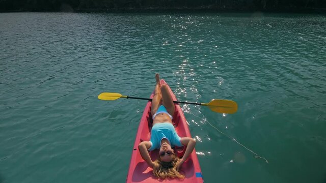 Pretty slim woman in elegant swimsuit lies in pink plastic kayak drifting on azure ocean at exotic resort upper view. Traveling to tropical countries. Girl is sailing on kayak in ocean, view above.
