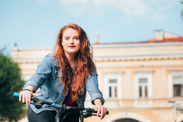 Fototapeta na wymiar Half-length portrait of long red curly hair caucasian teenager girl riding a modern bicycle in the old city center. Diverse people beauty or city bike hiring concept image.