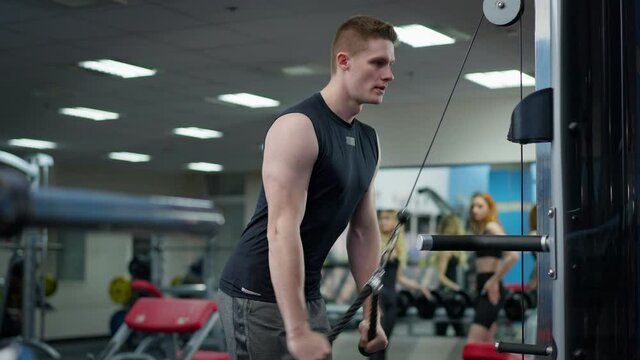 Concentrated fit young Caucasian sportsman training hand muscles using exercise machine in gym with blurred women talking at background. Handsome strong man working out indoors