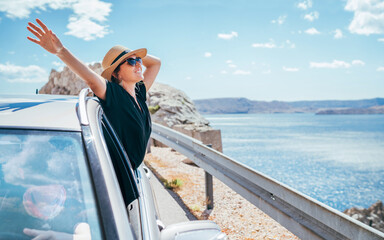 Cheerful Woman portrait enjoying the seaside road trip. Dressed a black dress, straw hat and sungllasses she wide opened arms and shining with happiness. Summer vacation traveling by auto concept.