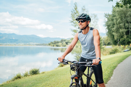 Portrait of a sincerely smiling man dressed in cycling clothes, helmet and sunglasses riding a bicycle on the asphalt out-of-town bicycle path along a mount lake. Active sporty people concept image.