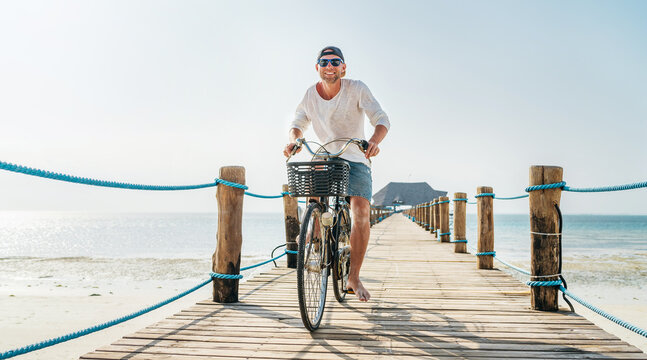 Portrait of a happy smiling man dressed in light summer clothes and sunglasses riding a bicycle on the wooden sea pier. Careless vacation in tropical countries concept image. Kiwengwa beach, Zanzibar