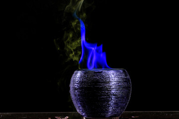 Pote de cerâmica, chama de fogo de cor azul e fumaça em fundo preto.  