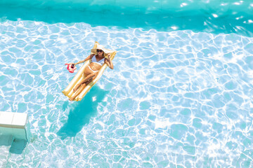 Top view of girl in a bathing suit and hat floating on inflatable gold mattress in the pool with drink cocktail. Summer holidays and relax concept