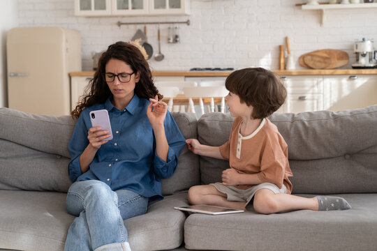Focused on mobile phone woman working mother pay no attention on bored small kid. Young mom or nanny texting in smartphone sitting on sofa next to little son need parent love care and communication