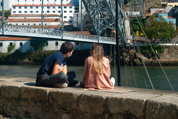 A couple is sitting at the Douro river embarkment talking, a bridge in the background - Portugal, Porto. - obrazy, fototapety, plakaty