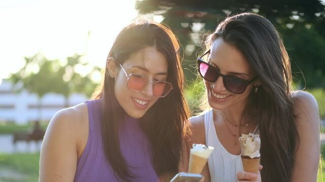 Video of  two beautiful young women eating ice cream while taking a photo with mobile phone in the park.