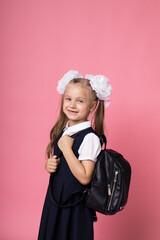 A European girl with white bows and a bag is happy to go to study. A schoolgirl on a pink background. Portrait of a smiling girl in a school uniform.