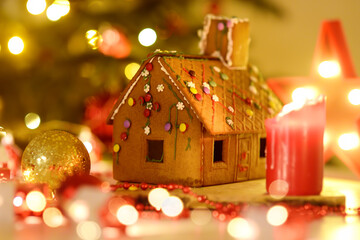Sweet homemade gingerbread house decorated candies and glazed is standing on table near Christmas tree with decorations lights, candles, holidays garlands.