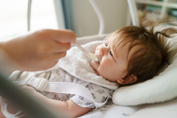 First meal side view small caucasian baby sitting or lying while hands of her unknown mother feeding her with plastic spoon containing organic rice cereal mash