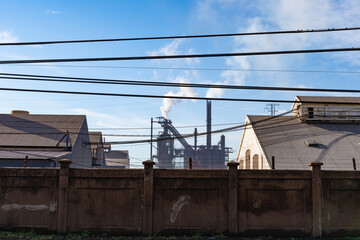 Steel mill blast furnaces behind a tall wall, smokestacks and smoke into blue skies, horizontal aspect