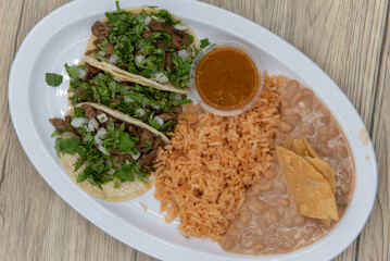 Overhead view of hearty plate of three loaded carne asada tacos with cilantro, served with rice and refried beans