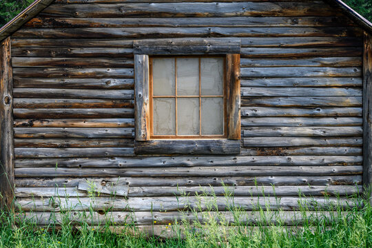 Rustic log cabin with wood frame window and tall grass in front, on a sunny day
