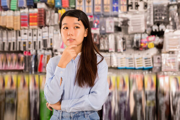 Portrait of cheerful Asian girl having doubts about choice of goods in cosmetics store