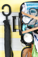 on a gray-yellow background, an open suitcase for traveling and a set of items for relaxing in it, a towel, an alarm clock, flip-flops, a diving mask, shorts, a closed umbrella next to it