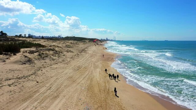 Aerial: Tourist Paragliding On Sunny Day By Coastline, Drone Flying Over People Walking At Beach During Sunny Day - Ashkelon, Israel