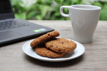 Pile of three delicious cookies on white plate. Blurry white glass and laptop on the background
