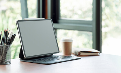Blank screen tablet with magic keyboard and supplies on wooden table in modern office room.