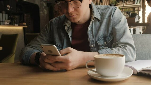 Close up of man's hand holding smart phone and typing with a thumb. He is at the cafe. Still shot, low angle, anonimous