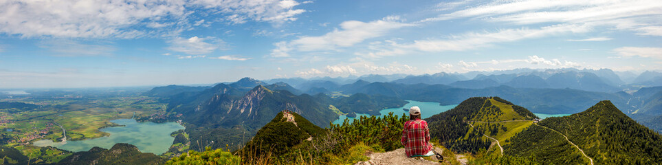 Obraz premium Woman sitting and relaxing at top of Mountain Herzogstand after hiking. looking at the spectacular bavarian Prealps mountains panorama with Lakes (Kochelsee, Walchensee) Europe, Germany