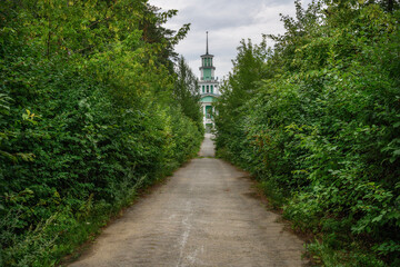 A green alley of deciduous shrubs, stretching into the distance and leading to an old building with a spire and a star. Linear perspective on a rural street in M. Istok (Ural, Russia). Summer day 