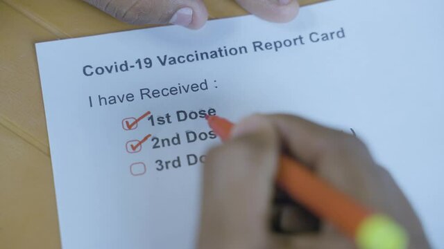 POV close up shot of hands checking Covid-19 vaccine report card and ticking 3rd or booster dose after vaccination.