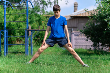 teenage boy exercising outdoors, sports ground in the yard, he squats and does a warm-up, healthy lifestyle