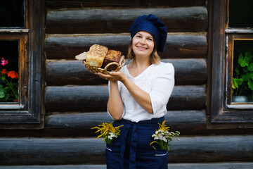 a smiling baker woman with a basket of bread in her hands in a chef's hat and apron against the background of a wooden log village house. Yeast-free wheat and rye bread with sourdough. Eco bakery.