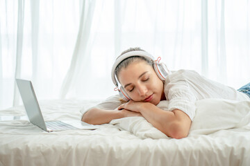 Lazy young student woman lying down on bed sleep with laptop computer listening to music with headphone at bedroom in morning . sleepy to learning online at home . take a nap